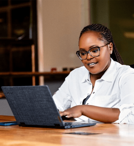 Mary Chege smiling while working on a laptop at a wooden table.